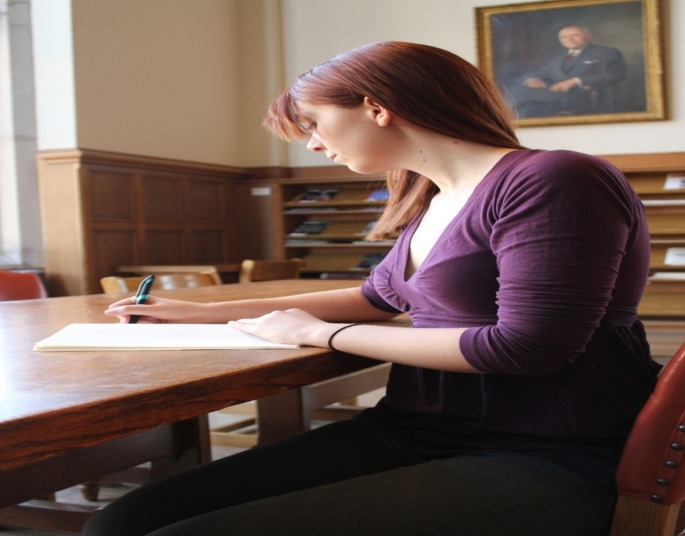 Das Bild zeigt eine junge Frau mit Papier und Stift an einem Schreibtisch sitzend.